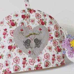Peut inclure: Un couvre-thé blanc avec un motif floral et en forme de cœur. Un patch en forme de cœur gris présente deux chérubins et des détails brodés. Un ruban vert et des accents floraux rouges complètent le design. Une théière blanche avec des fleurs est en arrière-plan.