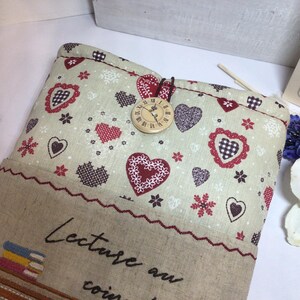 May include: A beige fabric book sleeve with a heart and floral pattern. The top flap features a wooden clock button closure. The bottom section has embroidered text and a decorative border. The overall theme is a cozy, romantic aesthetic.