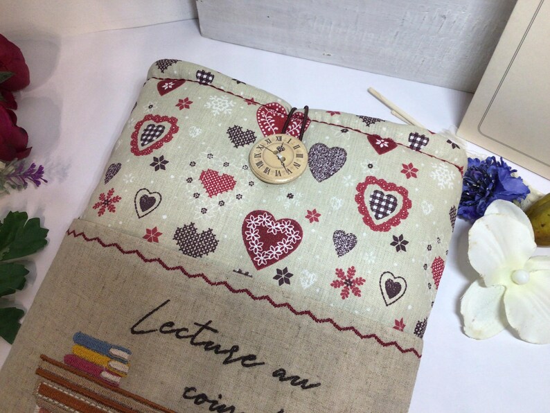 May include: A beige fabric book sleeve with a heart and floral pattern. The top flap features a wooden clock button closure. The bottom section has embroidered text and a decorative border. The overall theme is a cozy, romantic aesthetic.