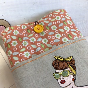 May include: A beige linen tote bag with a pocket featuring an embroidered image of a woman with brown hair, wearing sunglasses and a yellow headband. The tote bag has a floral print lining with orange, white, and green flowers.