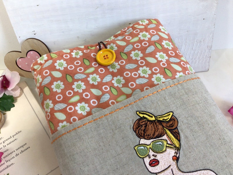 May include: A beige linen tote bag with a pocket featuring an embroidered image of a woman with brown hair, wearing sunglasses and a yellow headband. The tote bag has a floral print lining with orange, white, and green flowers.