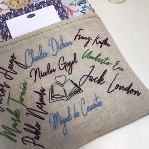 May include: A beige linen book sleeve with embroidered names of famous authors in various colors. The names include Charles Dickens, Franz Kafka, and Jack London. A notebook and clock are visible in the background.
