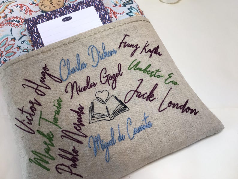 May include: A beige linen book sleeve with embroidered names of famous authors in various colors. The names include Charles Dickens, Franz Kafka, and Jack London. A notebook and clock are visible in the background.