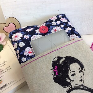 K&ouml;nnte beinhalten: Eine Buchh&uuml;lle aus Leinen mit einem Blumendruck und einem rosafarbenen Knopf. Die H&uuml;lle hat eine Tasche f&uuml;r ein Tablet oder einen E-Reader. Die Vorderseite der H&uuml;lle zeigt ein gesticktes Bild einer Frau mit schwarzen Haaren und einer rosa Blume.