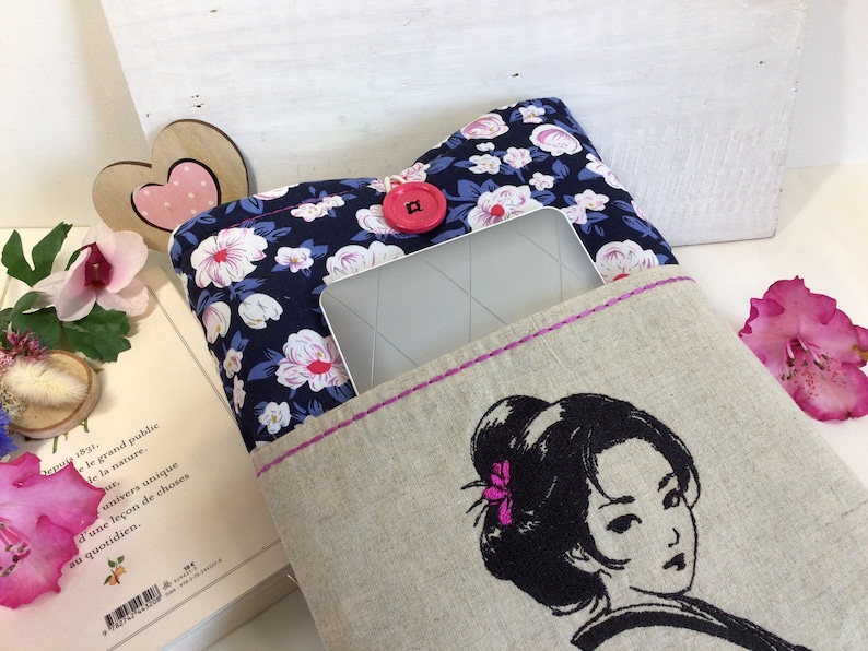K&ouml;nnte beinhalten: Eine Buchh&uuml;lle aus Leinen mit einem Blumendruck und einem rosafarbenen Knopf. Die H&uuml;lle hat eine Tasche f&uuml;r ein Tablet oder einen E-Reader. Die Vorderseite der H&uuml;lle zeigt ein gesticktes Bild einer Frau mit schwarzen Haaren und einer rosa Blume.