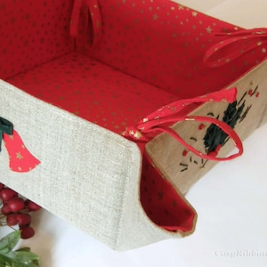 May include: A beige linen basket with red and gold fabric lining. The basket has two green bows with gold bells and red fabric accents. The basket has red fabric handles.