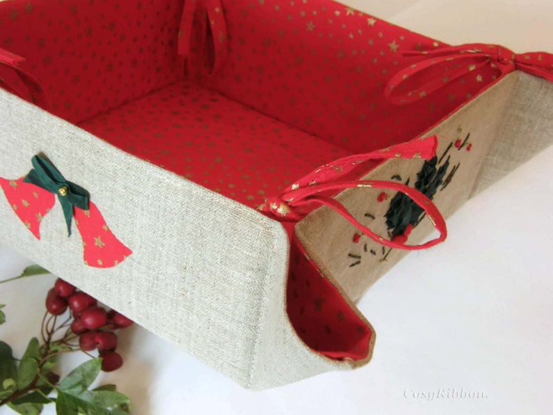 May include: A beige linen basket with red and gold fabric lining. The basket has two green bows with gold bells and red fabric accents. The basket has red fabric handles.