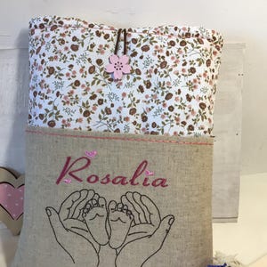 May include: A beige linen bag with a floral print top flap. The bag has a pink embroidered flower and the name "Rosalia" embroidered on the front. The bag also features two embroidered hands holding a pair of baby feet.