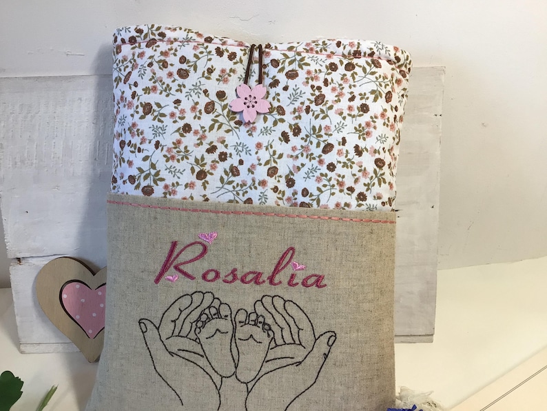 May include: A beige linen bag with a floral print top flap. The bag has a pink embroidered flower and the name "Rosalia" embroidered on the front. The bag also features two embroidered hands holding a pair of baby feet.