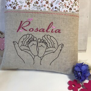 May include: A beige linen pillow with pink embroidery. The embroidery includes the name "Rosalia" and two hands holding a pair of baby feet.