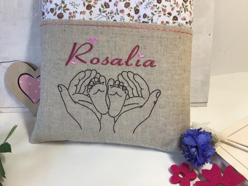 May include: A beige linen pillow with pink embroidery. The embroidery includes the name "Rosalia" and two hands holding a pair of baby feet.