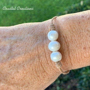 May include: A tan leather bracelet with three white pearls. The bracelet is tied with a knot on the wrist.