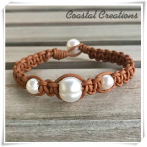 Puede incluir: Un brazalete de cuero marrón con un diseño trenzado y cuatro perlas blancas. El brazalete está sobre una superficie de madera. Coastal Creations está escrito en la esquina superior derecha de la imagen.