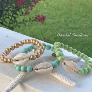 May include: Three bracelets with gold, light green, and turquoise beads. Each bracelet has a white shell charm and gold accents. The bracelets are on a white surface with a starfish.