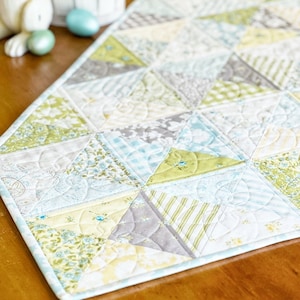 May include: A quilted table runner featuring a patchwork design in shades of blue, green, and white. The runner is crafted from a variety of fabrics with floral and geometric patterns.