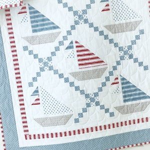 May include: A white nautical-themed quilt featuring sailboat designs. The sailboats have red and blue striped or star-patterned sails and gray hulls. The quilt has a red and white striped border and a blue patterned edge.