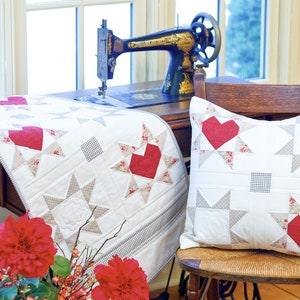 May include: A vintage sewing machine sits on a wooden table with a white and red quilt and a pillow with a red heart design. A bouquet of red flowers is in a metal vase on the table.
