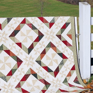 Pode incluir: Uma colcha com um padrão geométrico de triângulos vermelhos, verdes e marrons sobre um fundo branco. A colcha está dobrada e drapeada sobre um poste de cerca branco. A colcha está em um campo com grama verde e folhas marrons.