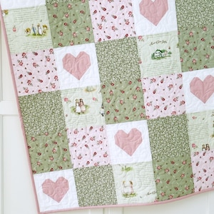 May include: A patchwork quilt featuring a repeating pattern of pink hearts, floral prints, and scenes of people and houses. The quilt has a white background with a pink and white gingham border. The quilt is made of squares of fabric.