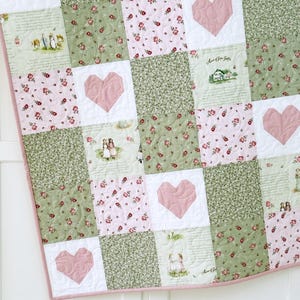 May include: A patchwork quilt with a repeating pattern of pink hearts, floral prints, and scenes of people and houses. The quilt has a pink and white gingham border and is made of various fabrics in shades of pink, green, and white.