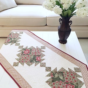 May include: A white table runner with a red and green floral pattern. The runner has a diamond-shaped design with a border of red and white. The runner is on a white table with a vase of white flowers.