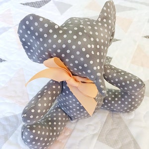 Puede incluir: Un elefante de peluche gris y blanco con lunares y un lazo de color melocotón. El elefante está sentado en una colcha blanca y gris.