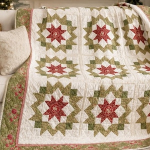 May include: A quilted throw blanket featuring a repeating star pattern in shades of red, olive green, and cream. The quilt has a decorative border with a floral motif and a small red and white checkered trim, draped over a white sofa.