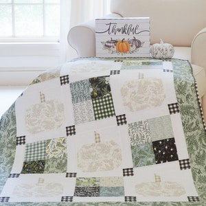 May include: A white quilt with a green and black checkered border. The quilt is made up of squares with different patterns, including a floral pattern, a plaid pattern, and a solid green pattern. The quilt features a design of pumpkins with a stem and leaves. The quilt is sitting on a white couch with a pillow and a wooden sign that says "Thankful" with a pumpkin design.