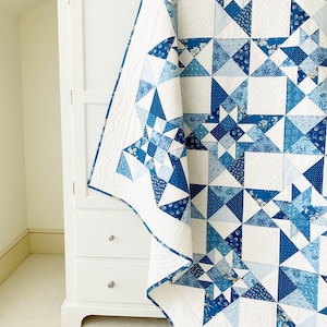 Peut inclure: Une courtepointe blanche et bleue avec un motif g&eacute;om&eacute;trique d'&eacute;toiles et de carr&eacute;s. La courtepointe est pli&eacute;e et drap&eacute;e sur un meuble blanc avec des tiroirs.