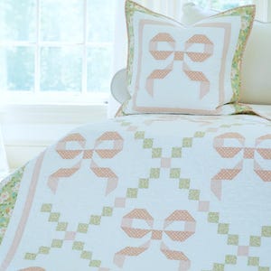 May include: A white quilt and pillow set with a repeating bow pattern in peach and white polka dots. The quilt has a floral border and a matching pillow. The design is set against a soft, light-filled background.