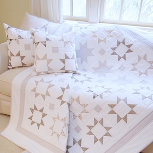 Puede incluir: Una colcha blanca y beige con un patrón de estrellas. La colcha está doblada y drapeada sobre un sofá blanco con dos fundas de almohada a juego.