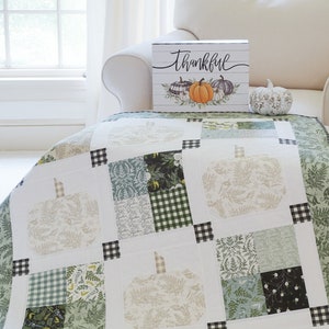 May include: A patchwork quilt featuring a repeating pattern of fabric pumpkins in shades of sage green, cream, and white, accented with black and white gingham squares. A rectangular sign with the word Thankful and a pumpkin illustration sits nearby.