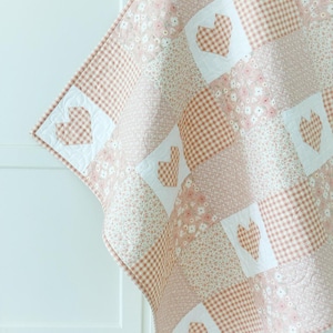 May include: A pink and white patchwork quilt with a heart pattern. The quilt is made of different fabrics, including gingham and floral prints.