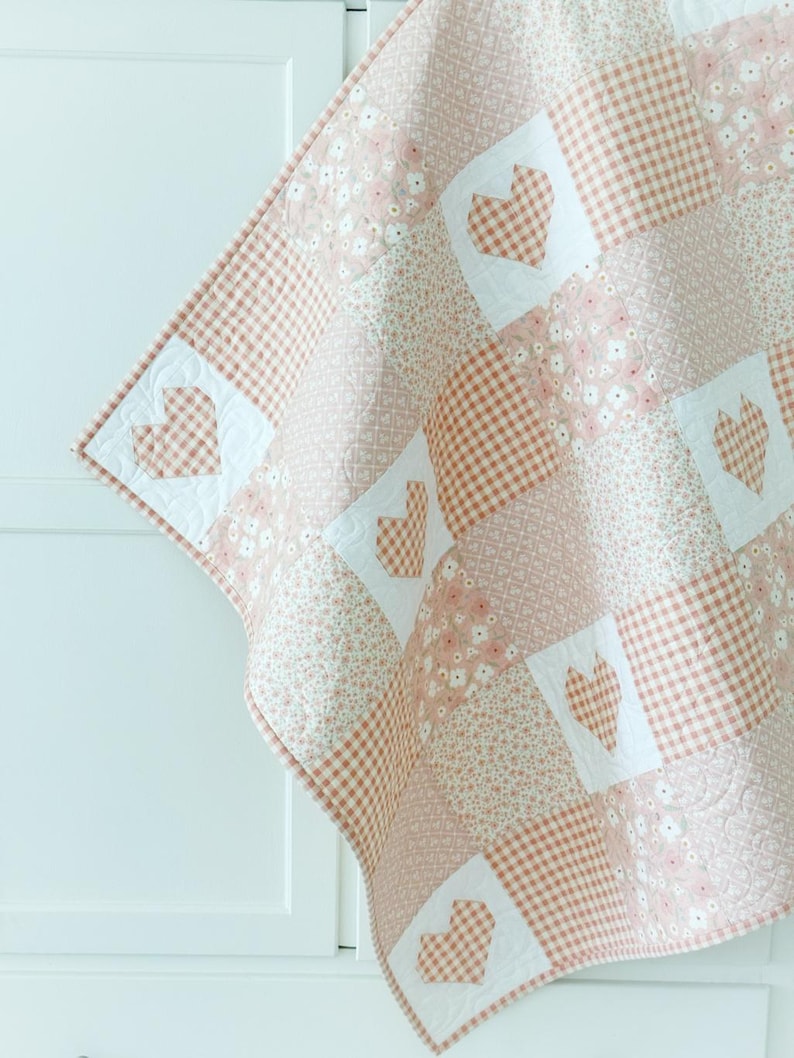 May include: A pink and white patchwork quilt with a heart pattern. The quilt is made of different fabrics, including gingham and floral prints.