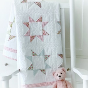 May include: A white baby quilt with a pink and green floral pattern and a pink and white gingham border. The quilt is folded and draped over a white rocking chair with a pink teddy bear sitting in front of it.