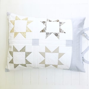 Puede incluir: Una almohada blanca con un borde gris y cuatro parches en forma de estrella en diferentes tonos de beige y marrón. La almohada tiene una corbata de tela gris.
