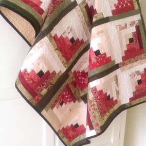Peut inclure: Une courtepointe rouge, verte et cr&egrave;me avec un motif de cabane en rondins. La courtepointe est pli&eacute;e et drap&eacute;e sur une commode blanche avec des tiroirs.
