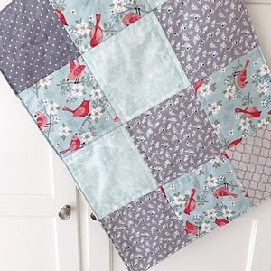 May include: A baby quilt with a patchwork design featuring a variety of fabrics. The quilt is made up of squares with different patterns, including a floral pattern with red birds, a gray polka dot pattern, and a solid light blue pattern. The quilt is perfect for a nursery or a child's bedroom.