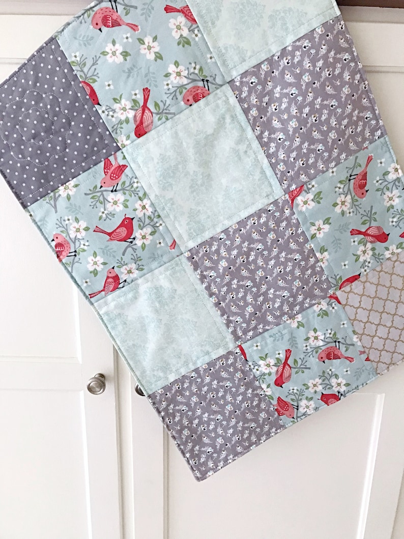May include: A baby quilt with a patchwork design featuring a variety of fabrics. The quilt is made up of squares with different patterns, including a floral pattern with red birds, a gray polka dot pattern, and a solid light blue pattern. The quilt is perfect for a nursery or a child's bedroom.