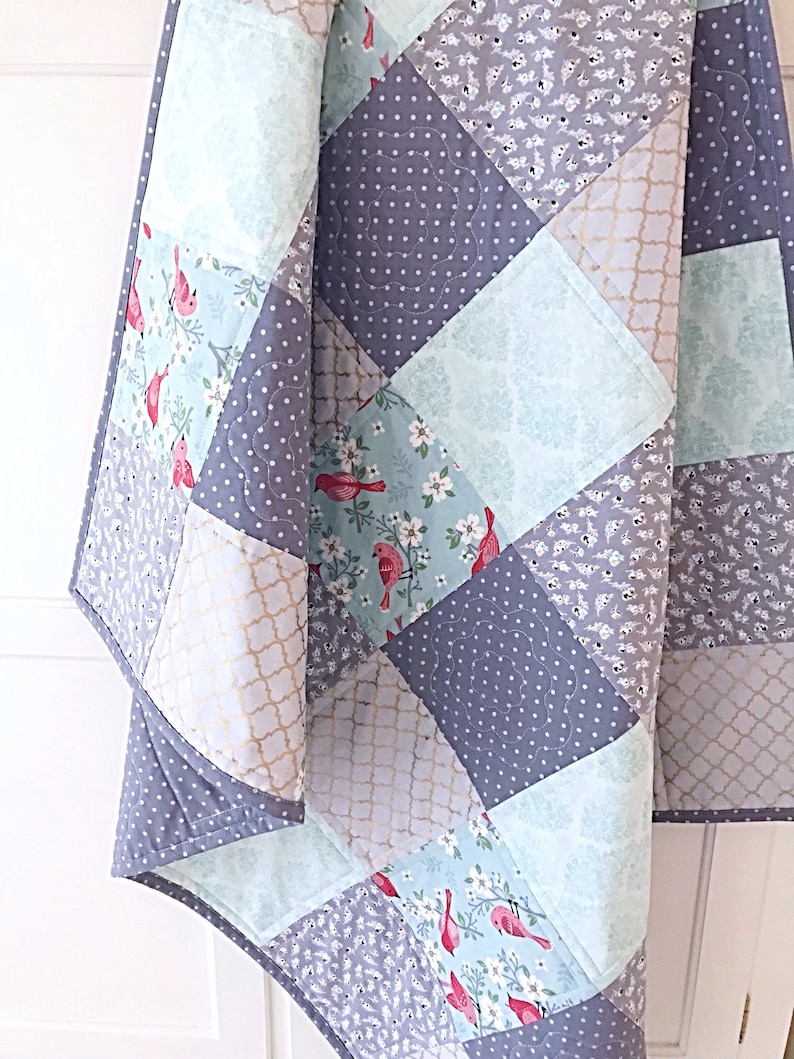 May include: A quilt made of patchwork squares in shades of gray, light blue, and gold. The squares feature different patterns, including polka dots, floral designs, and geometric shapes. There are also red birds on some of the squares.