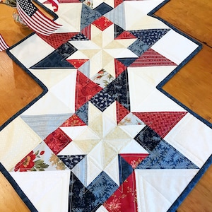 May include: A red, white, and blue quilted table runner with a star pattern. The runner is made of various fabrics with floral and geometric designs.