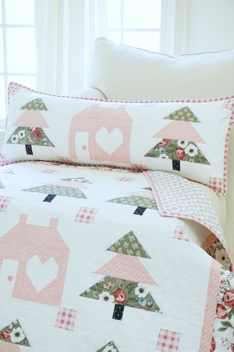 Puede incluir: Una colcha blanca con un dise&ntilde;o de patchwork rosa y verde con casas y &aacute;rboles. La colcha est&aacute; sobre una cama blanca con una almohada a juego.