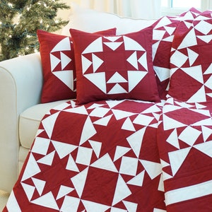 Pode incluir: Uma colcha vermelha e branca com um padrão de estrelas. A colcha está em um sofá branco com duas almofadas vermelhas e brancas. Uma árvore de Natal está no fundo.