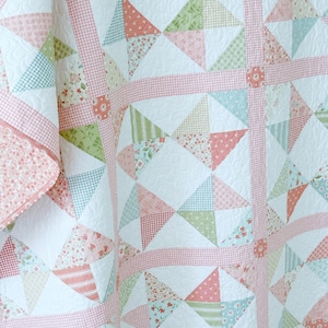 Könnte beinhalten: Eine handgefertigte Steppdecke mit Patchwork-Design. Die Steppdecke hat einen weißen Hintergrund mit geometrischen Formen in Rosa-, Grün- und Blautönen. Die Steppdecke ist mit einem rosa Vichy-Muster eingefasst und hat eine gesteppte Textur.