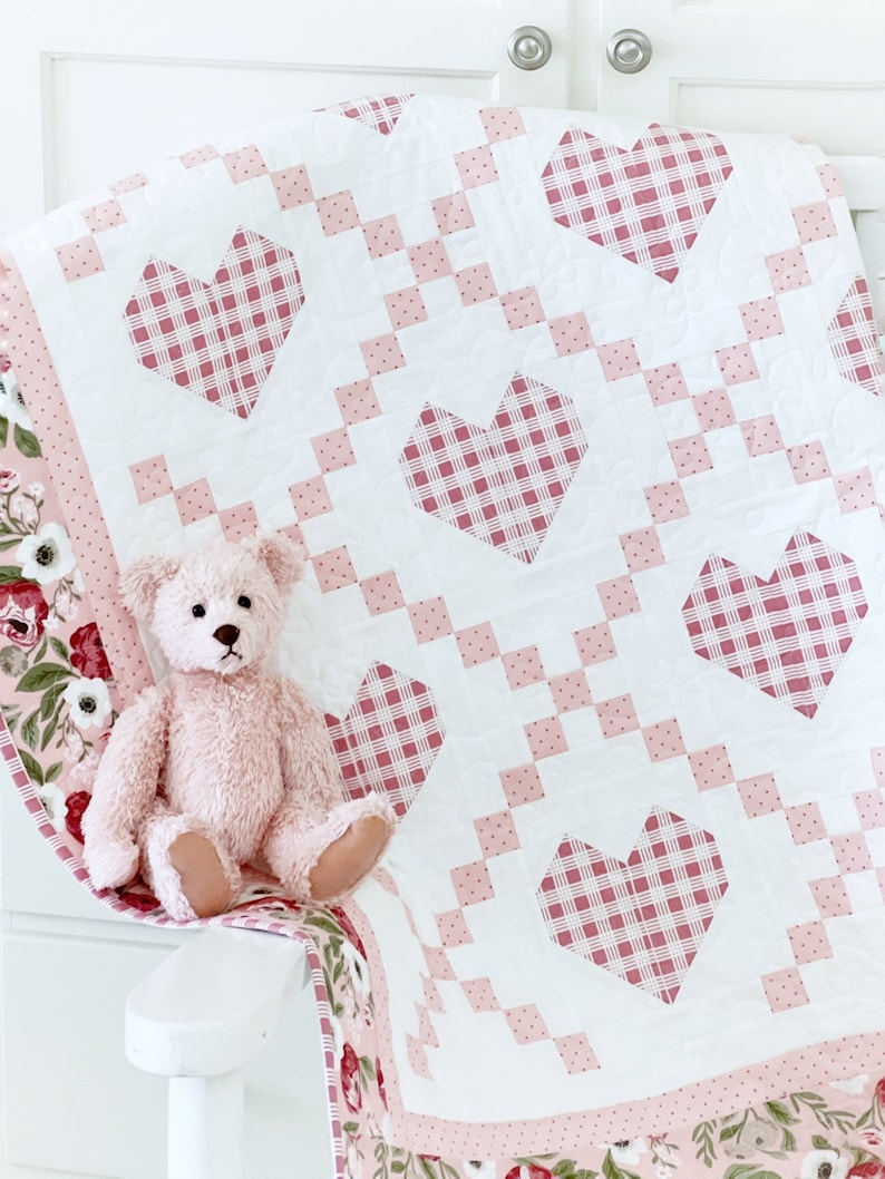 May include: A pink and white quilt with a checkered heart pattern. The quilt is folded and draped over a white chair with a pink teddy bear sitting on it.