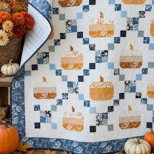 May include: A quilt featuring a repeating pattern of orange and white pumpkin shapes arranged in a grid. The design incorporates blue and white floral and geometric fabric squares, with a dark blue floral border and white quilted background.