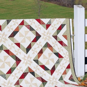 Puede incluir: Una colcha con un patrón geométrico en tonos rojos, verdes y beige. La colcha está drapeada sobre una valla blanca con un fondo de una colina verde cubierta de hierba y árboles.