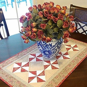 Op de afbeelding: Een tafelkleed met een wit en rood windmolenpatroon, een blauwe en witte porseleinen vaas met een bloemenpatroon en een boeket oranje rozen.