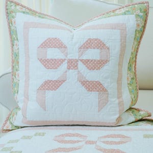 May include: A white and pink quilted pillow with a bow design. The bow is made of pink fabric with white polka dots. The pillow has a floral border and is on a white surface.
