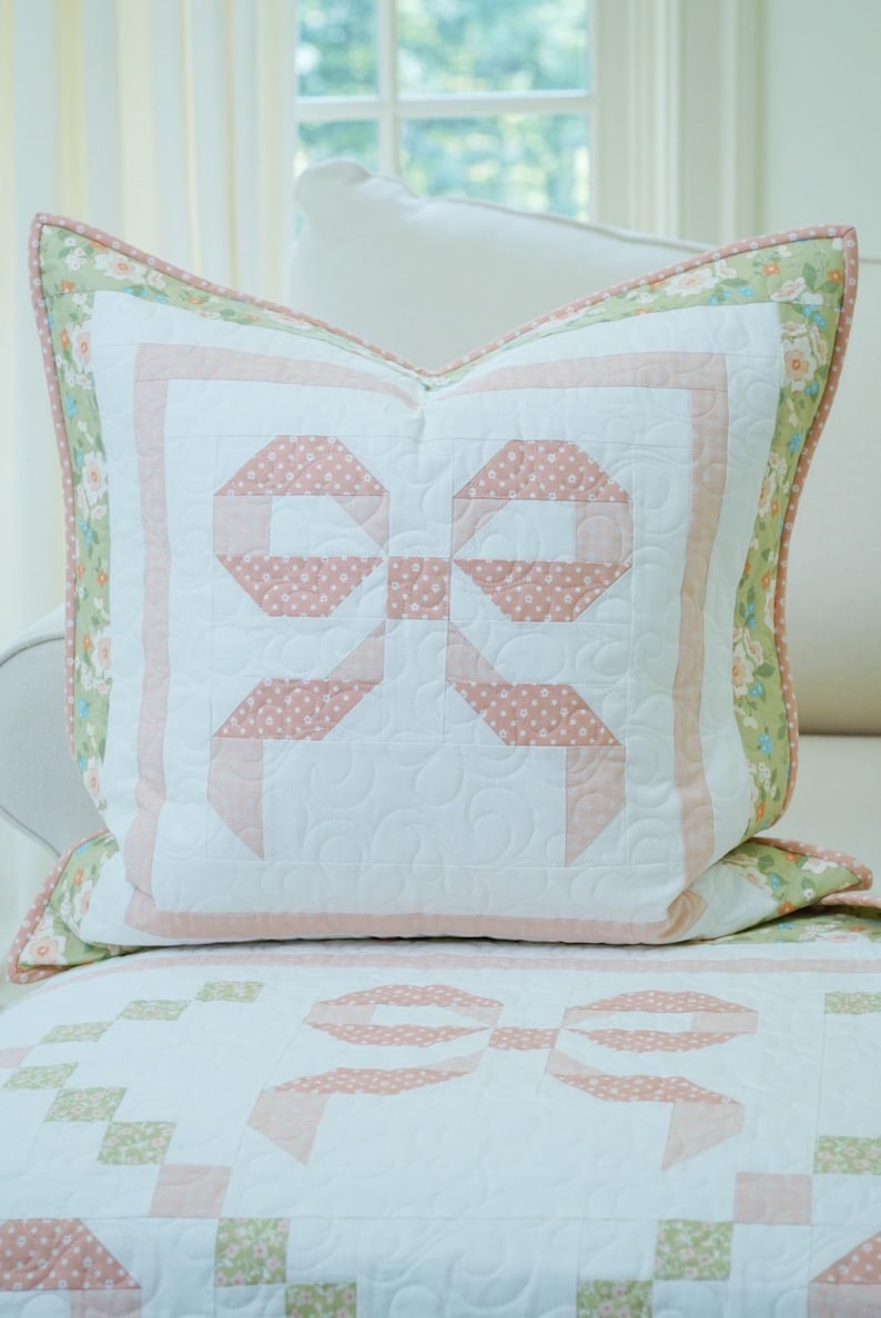 May include: A white and pink quilted pillow with a bow design. The bow is made of pink fabric with white polka dots. The pillow has a floral border and is on a white surface.